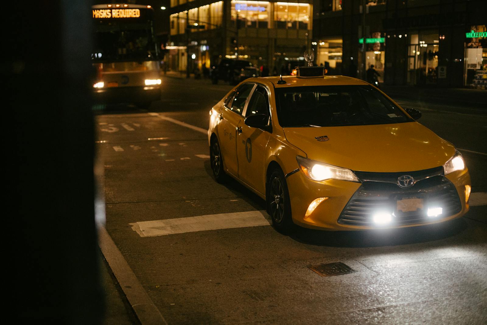 A yellow taxi cab waits at a city street intersection at night, illuminated by headlights.